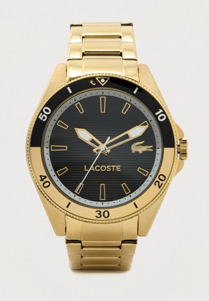 Złoty metalowy zegarek na rękę z czarną tarczą, złotymi znacznikami godzin i wskazówkami, białą wskazówką sekundową oraz logo Lacoste na tarczy.