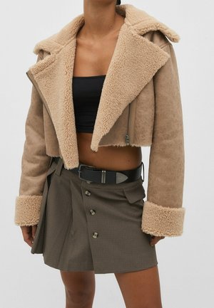 Femme portant une veste courte en peau retournée beige, un crop top noir et une jupe marron taille haute boutonnée avec une ceinture noire.
