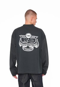 Cămașă neagră cu mâneci lungi, având un design grafic alb care prezintă un insigne cu "Multiply Apparel 2025" și "Made for the Thrill."