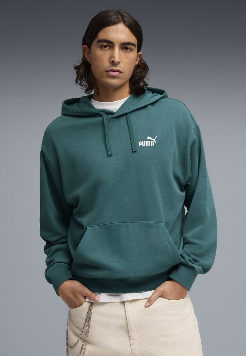 Ung person med axellångt hår som bär en teal Puma-hoodie och ljusfärgade byxor med händerna i fickorna mot en grå bakgrund.