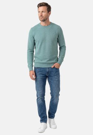 Mann trägt einen helltürkisfarbenen Rundhalspullover, blaue Jeans und weiße Turnschuhe, steht mit einer Hand in der Tasche und schaut nach links.