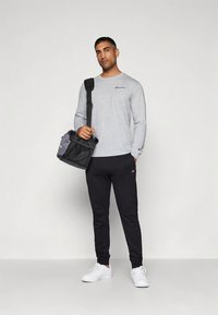 Gray long-sleeve shirt, black sweatpants, and white sneakers. The individual holds a black and gray duffel bag. Logo on shirt and pants.