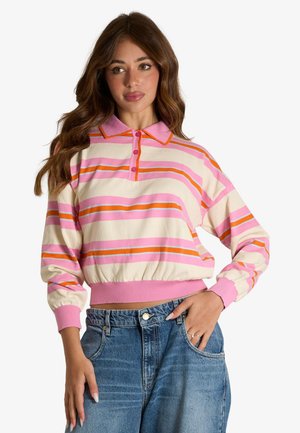 Donna con lunghi capelli castani che indossa un maglione polo a righe rosa, arancione e crema a maniche lunghe e jeans blu con la mano in tasca.