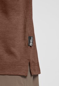 T-shirt en coton marron avec fente sur le côté et ourlet cousu. Inclut une étiquette noire avec le logo "Jack Wolfskin" et un motif de patte.