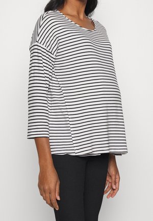 Femme enceinte portant un haut de maternité à manches longues rayé blanc et noir et un pantalon noir, visible des épaules aux hanches.