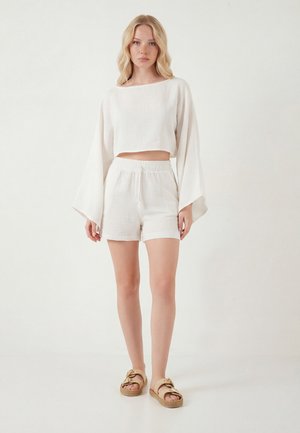 Kobieta w luźnej, białej, długorękawowej krótkej bluzce i pasujących szortach, stojąca boso na białym tle.