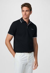 Polo negra con cuello y detalles de manga en blanco, con un pequeño logo en el pecho. Combinada con pantalones de color claro.