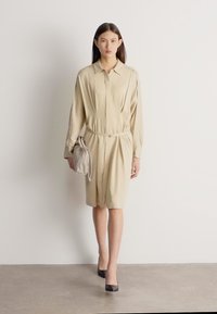 Žena v béžovom košeľovom šatách so zápasom opaskom, nesie zladenú kabelku, kráča po sivom podlaží pred jednoduchou bielou stenou.