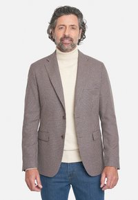 Blazer marrón de patrón de espiga, de un solo botón con solapas de pico y dos bolsillos delanteros, llevado sobre un suéter de cuello alto crema y jeans azules.