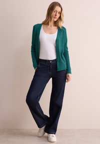 Tjalfarbige, gerippte Strickjacke mit Knöpfen vorne, getragen über einem weißen Tanktop; kombiniert mit dunkelblauen, weit geschnittenen Jeans und weißen Sneakers. Minimalistisches Design.