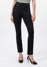 Slim-fit černé kalhoty s zlatými knoflíky na přední straně, nošeny s černými vysokými podpatky a zasunutým šedým topem.