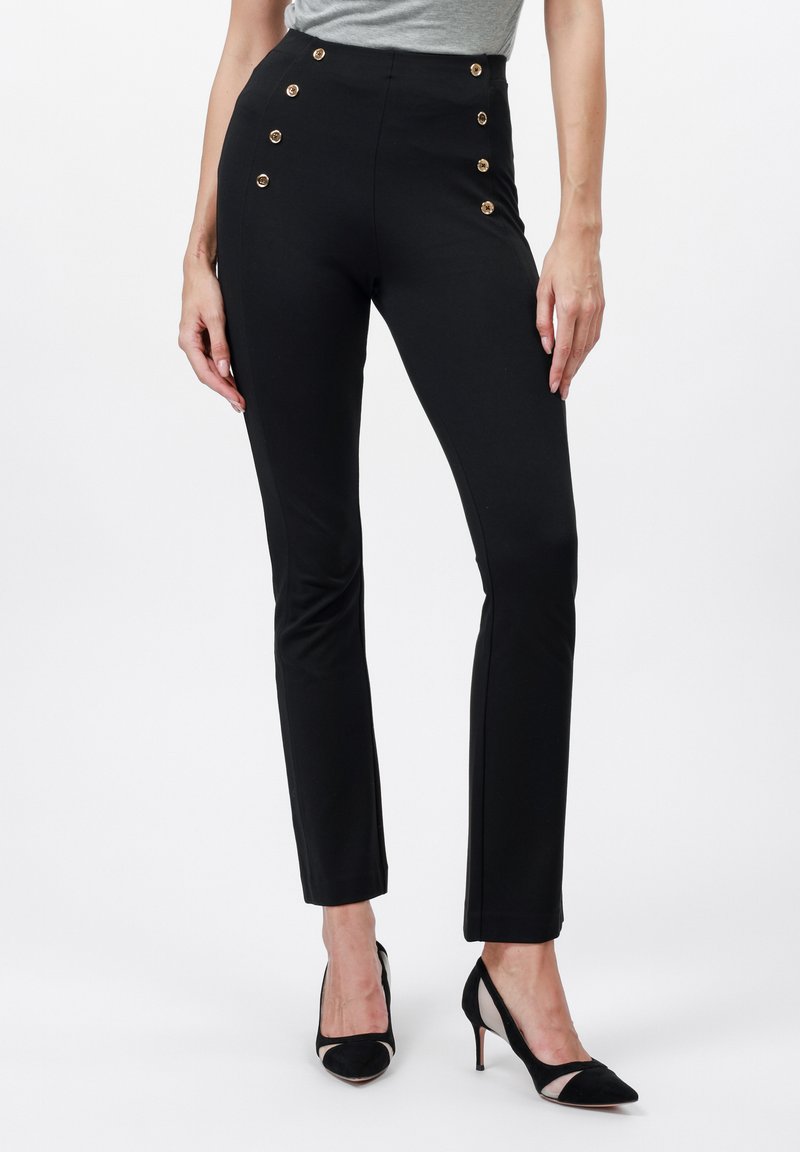 Slim-fit černé kalhoty s zlatými knoflíky na přední straně, nošeny s černými vysokými podpatky a zasunutým šedým topem.