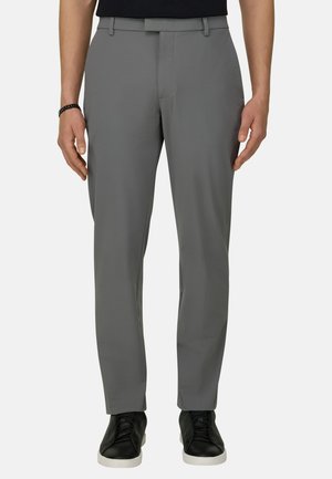 Mann trägt gerade geschnittene graue Hose, schwarzen Gürtel, schwarze Sneakers mit weißen Sohlen, steht vor einem schlichten Hintergrund.