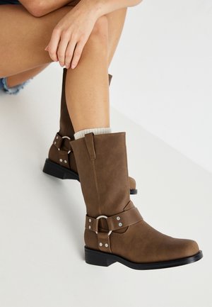 Personne portant des bottes en cuir marron à mi-mollet avec des détails de boucles argentées et des chaussettes de couleur claire, assise avec une jambe croisée sur l'autre.