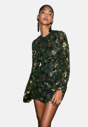 Woman wearing a dark green long-sleeve mini dress with gold and black floral sequins, holding a clutch, and wearing long black tassel earrings.