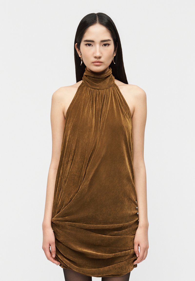 Γυναίκα με μακριά σκούρα μαλλιά, ντυμένη με ένα καφέ βελούδινο φόρεμα χωρίς μανίκια και με ψηλό λαιμό, με ρίγες υφάσματος και μικρά ασημένια σκουλαρίκια.