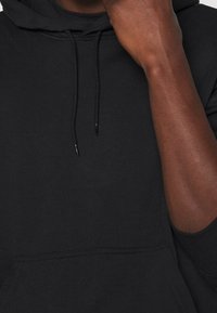 Zblízka záběr osoby, která má na sobě černou mikinu s kapucí a šňůrami, částečně si zakrývá ústa rukou.