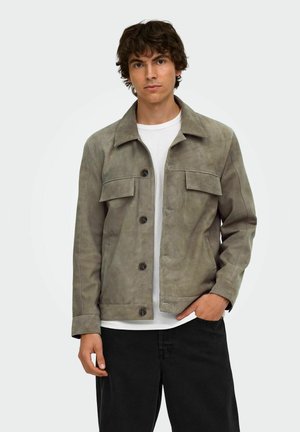 Jeune homme portant une veste grise boutonnée sur une chemise blanche avec un pantalon noir, debout devant un fond clair uni.