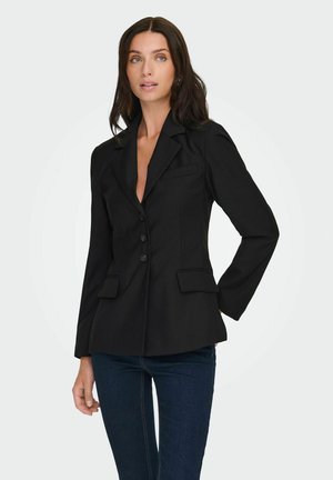 Femme aux cheveux longs bruns portant un blazer noir cintré à trois boutons et un jean bleu foncé, debout devant un fond clair.