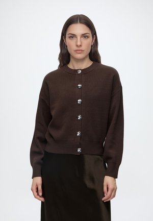 Femme aux cheveux bruns lisses portant un cardigan boutonné brun foncé avec des boutons argentés et une jupe noire sur un fond blanc uni.