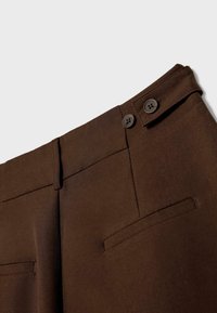 Gros plan d'un pantalon marron sur mesure avec patte de bouton latérale et poches passepoilées sur fond clair.