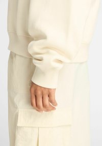 Sweatshirt couleur crème avec des poignets côtelés, associé à un tissu clair avec une poche et un design plissé. Texture douce visible.