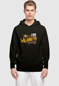 Černá mikina s kapucí a přední kapsou, která obsahuje grafický design se slovem "Hogwarts" napsaným žlutě na texturovaném pozadí s razítky.