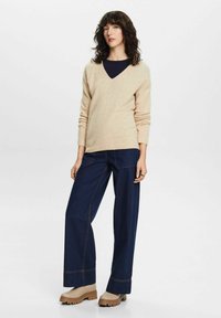 Beige V-hals trui in gebreide textuur, gecombineerd met donkerblauwe wijde denim jeans met contrasterende stiksels en tan enkellaarsjes.