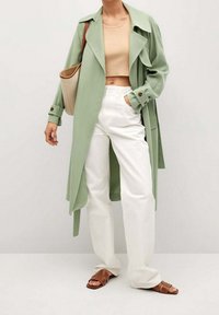 Žena v světle zeleném trenčkotě, béžovém cropped topu, bílých kalhotách, hnědých sandálech a s béžovou kabelkou přes rameno.