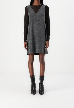 Personne debout portant une robe grise texturée sans manches, longueur genou, par-dessus un haut noir transparent à manches longues et des bottines noires.