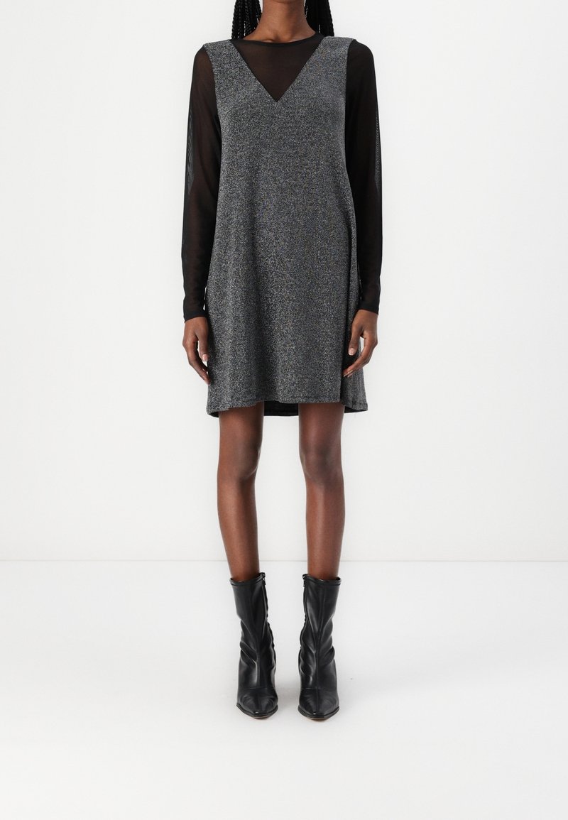 Personne debout portant une robe grise texturée sans manches, longueur genou, par-dessus un haut noir transparent à manches longues et des bottines noires.