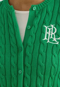 Strikket grønn cardigan med kabelmønster, ribbet krage og knapper, med en hvit brodert logo på venstre side.