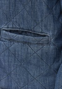 Detalj na jakni od kviltanog trapera koja prikazuje mali otvoreni džep i teksturiranu plavu tkaninu s dijamantnim uzorcima i kontrastnim šavovima.
