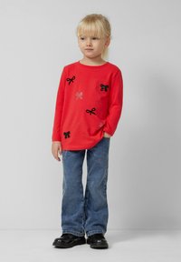 Rode lange mouwen shirt met zwarte strik applicaties en rhinestone accenten, gecombineerd met blauwe denim jeans en zwarte schoenen. Eenvoudige achtergrond.