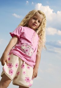 Lichtroze t-shirt met gerimpelde mouwen, met een cameragrafiek, gecombineerd met crèmekleurige shorts versierd met roze bloemmotieven.