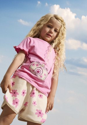 Lichtroze t-shirt met gerimpelde mouwen, met een cameragrafiek, gecombineerd met crèmekleurige shorts versierd met roze bloemmotieven.