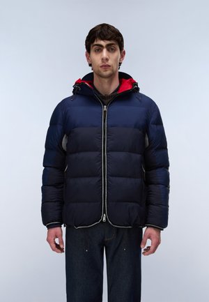 Giovane uomo in piedi che indossa una giacca imbottita blu navy e nera con fodera rossa nel cappuccio, abbinata a jeans blu scuro contro uno sfondo chiaro e semplice.