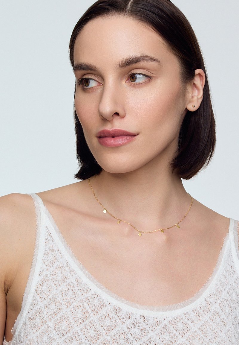 Kobieta z krótkimi ciemnymi włosami nosi delikatny złoty naszyjnik i lekką białą bluzkę, patrząc lekko na bok na jasnym tle.