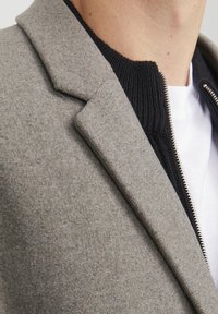Sivi jakna od vune s dodatkom poliestera, s reverom, teksturiranom tkaninom i crnim rebrastim pletenim visokim ovratnikom. Vidljiv srebrni detalj sa zatvaračem duž strane.