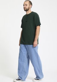 Mørkegrøn bomulds t-shirt med korte ærmer og broderet logo, parret med lyseblå bredbenede denimjeans og sorte sneakers.