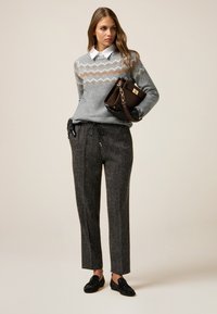 Suéter gris con un diseño estampado, camisa blanca con cuello debajo, combinado con pantalones oscuros gris oscuro a medida y mocasines negros. Bolso marrón en la mano.