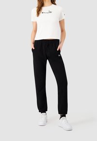 Valkoinen lyhythihainen t-paita mustalla logolla, yhdistettynä mustiin löysiin housuihin, joissa on joustava vyötärö ja pieni logo lantiolla. Valkoiset lenkkarit.