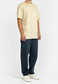 Beige bomullströja med rund halsringning och korta ärmar, med ett litet grönt kaktusmotiv på vänster bröst. Marinblå byxor.