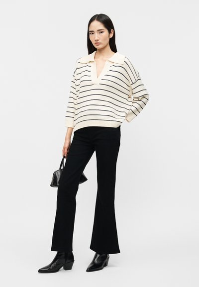 Γυναίκα που φοράει πουλόβερ με κρεμ και μαύρες ρίγες, μαύρο καμπάνα παντελόνι, μαύρα μπότακια αστραγάλου, κρατώντας μια μικρή μαύρη τσάντα, στέκεται μπροστά σε έναν απλό φόντο.