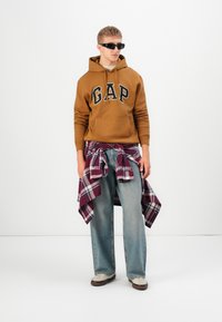 Haut à capuche marron avec le logo "GAP", chemise en flanelle à carreaux rouge et blanc nouée autour de la taille, jeans larges bleu clair, chaussures beiges et lunettes de soleil noires.