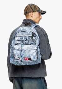 Denim-patterned backpack in light blue with multiple zippered pockets, adjustable straps, and a logo label at the bottom front.