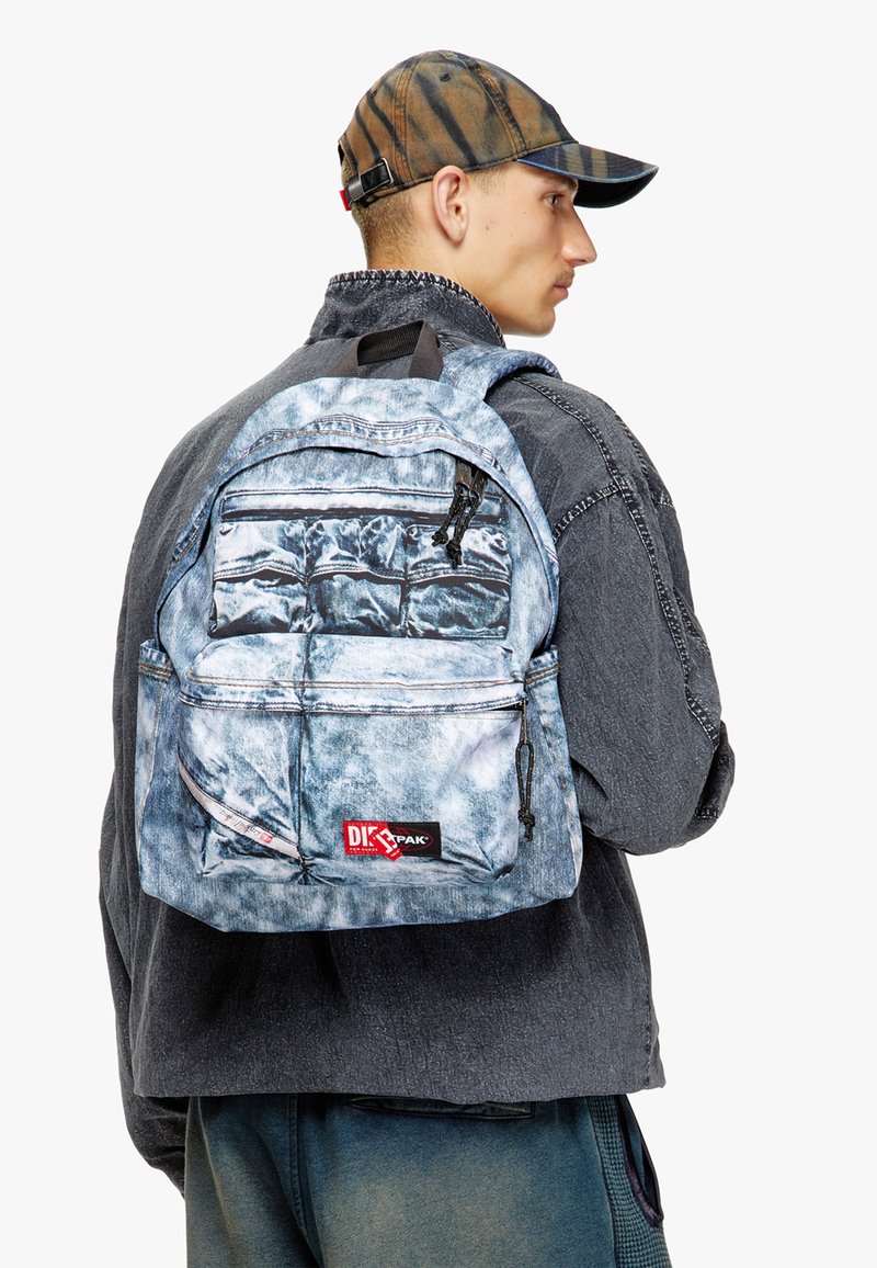 Denim-patterned backpack in light blue with multiple zippered pockets, adjustable straps, and a logo label at the bottom front.