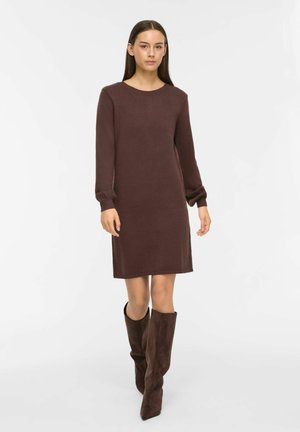 Femme en robe marron tricotée à manches longues et bottes marron en daim montantes jusqu'aux genoux, debout devant un fond blanc.