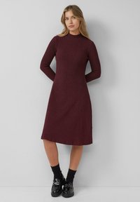 Burgundry gestricktes Kleid mit Stehkragen, langen Ärmeln und Midi-L�änge. Es hat ein strukturiertes Muster und wird mit schwarzen Schuhen und Socken kombiniert.