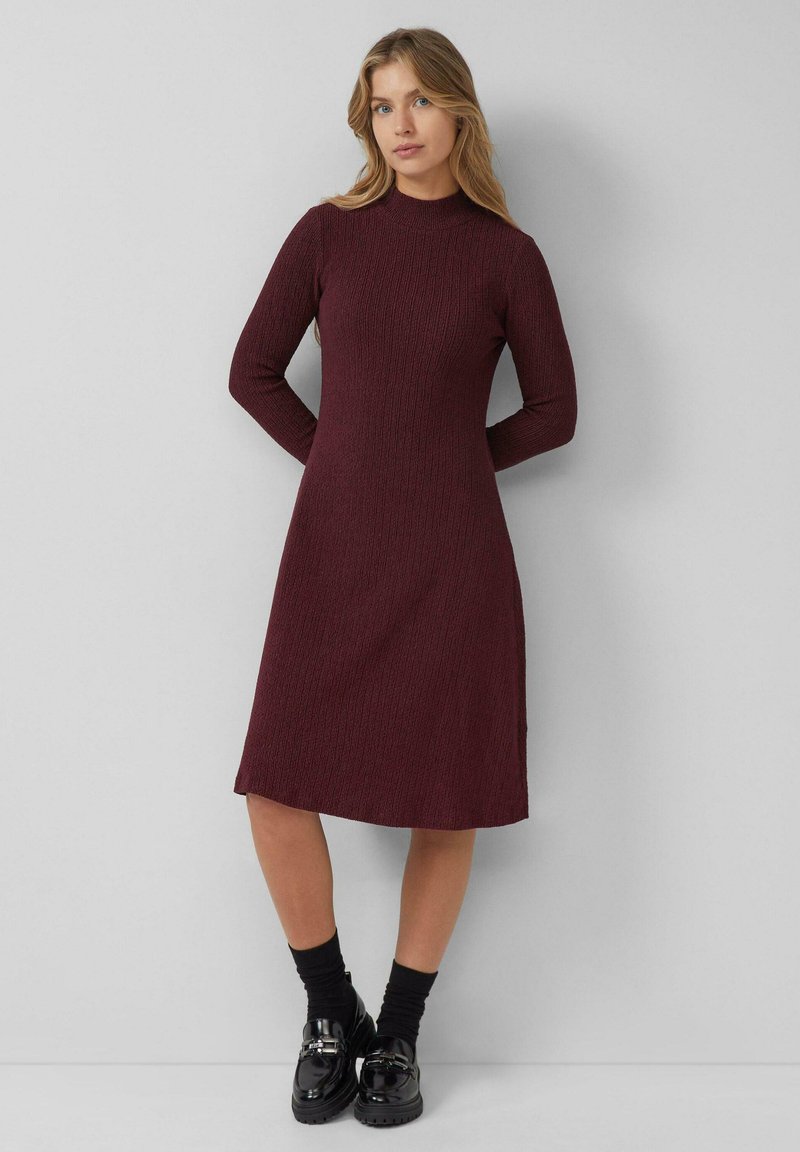 Burgundry gestricktes Kleid mit Stehkragen, langen Ärmeln und Midi-Länge. Es hat ein strukturiertes Muster und wird mit schwarzen Schuhen und Socken kombiniert.